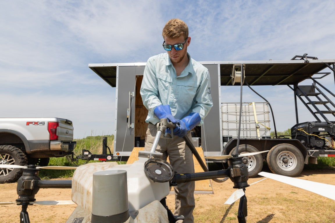 Person working on drone equipment outdoors with a truck and trailer in the background.
